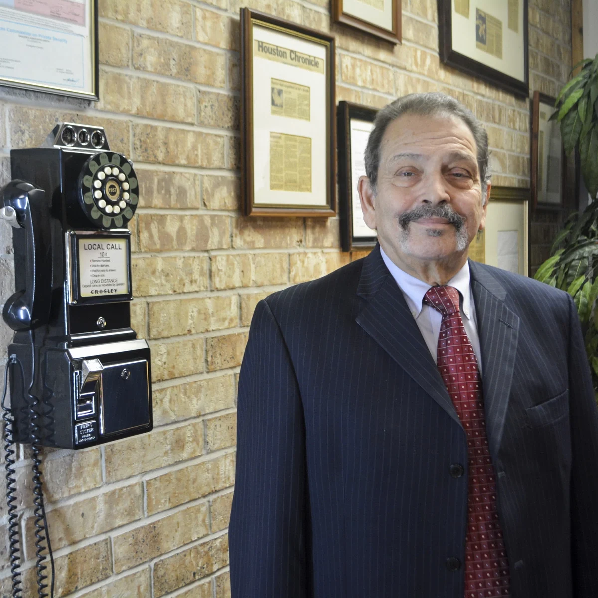 J.J. Gradoni in suit standing by brick wall with framed articles and vintage rotary phone