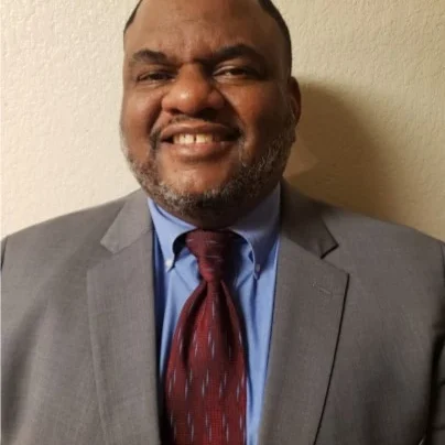 Will Hodge smiling in a gray suit and maroon tie for a professional headshot